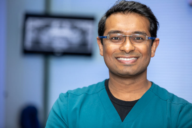 Smiling dentist wearing glasses and teal scrubs standing in a modern dental office with an X-ray monitor in the background.